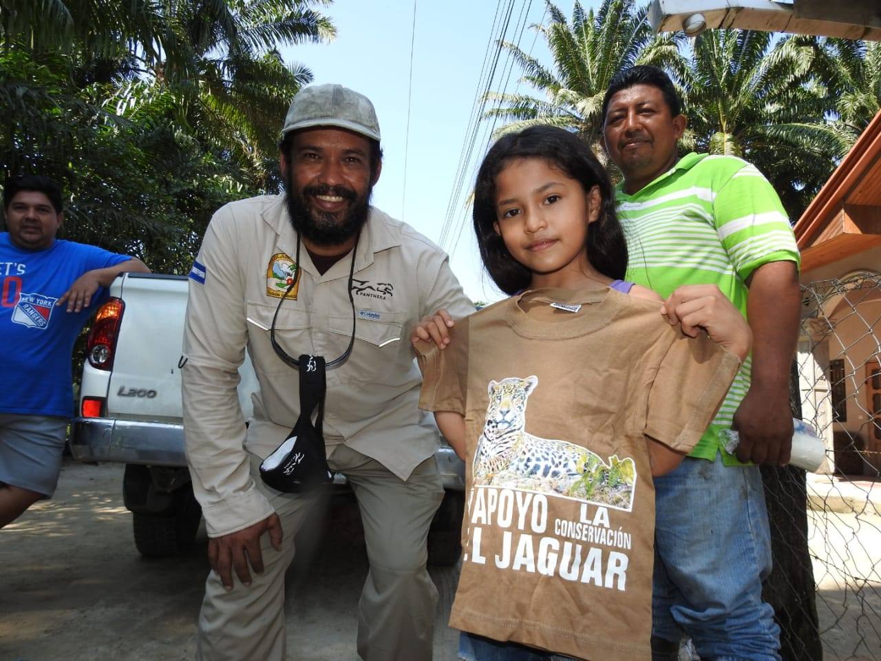 "Little girl holding t-shirt smiling at camera with three men in the background"